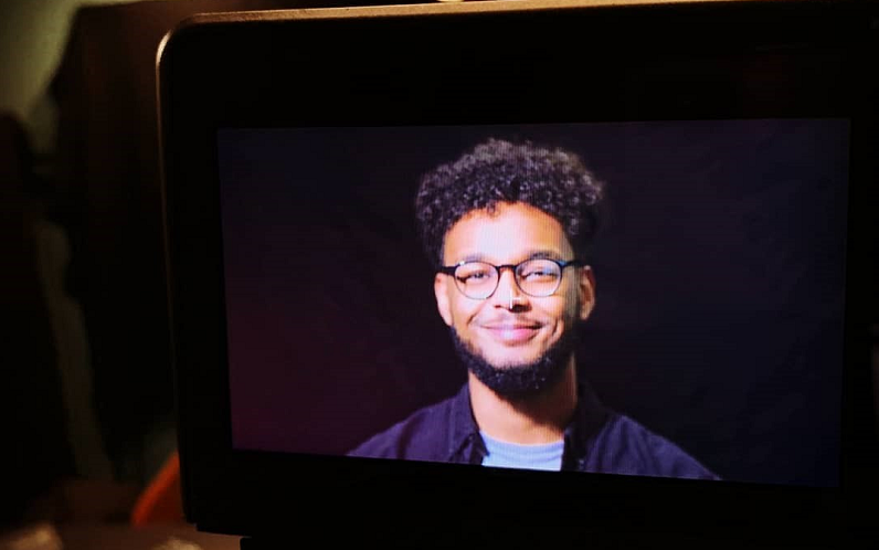 A picture of a camera where you can see Ahmed being filmed. He's got a big grin while standing in front of a black background. He's looking straight down the camera lens. He's wearing glasses and a blue t-shirt and a blue shirt,