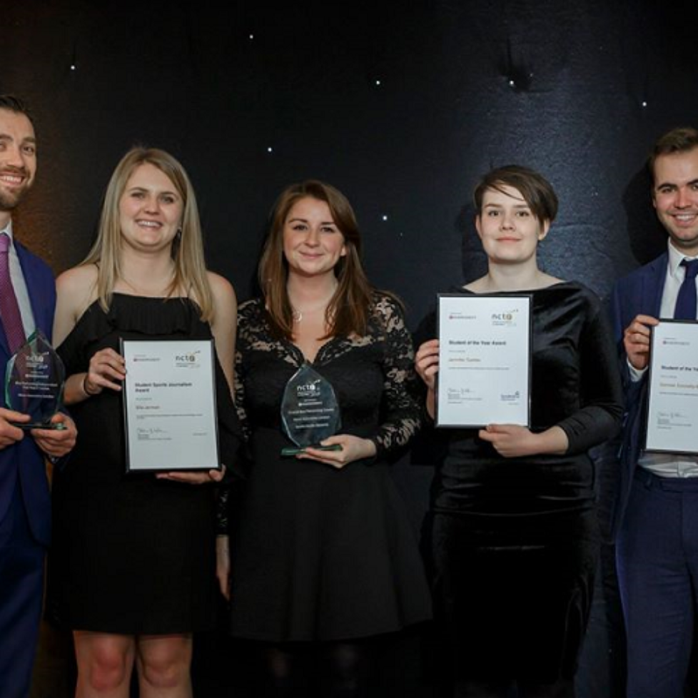 News Associates staff and trainees with their awards at the 2019 NCTJ Awards for Excellence. Graham Moody, Ella Jerman, Lucy Dyer, Jen Tombs and Cormac Connelly are standing on a stage in front of a black backdrop. They all look very smart. Lucy and Graham are holding glass awards while the trainees are holding framed certificates.