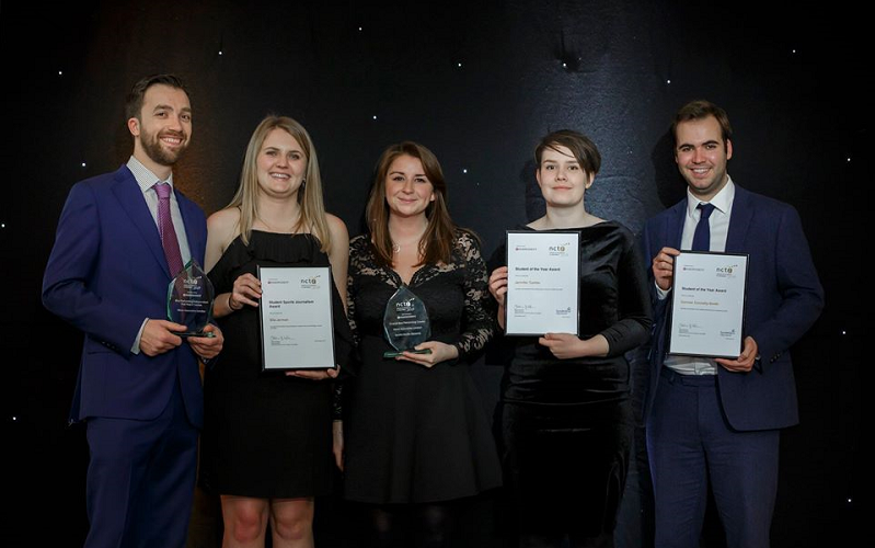 News Associates staff and trainees with their awards at the 2019 NCTJ Awards for Excellence. Graham Moody, Ella Jerman, Lucy Dyer, Jen Tombs and Cormac Connelly are standing on a stage in front of a black backdrop. They all look very smart. Lucy and Graham are holding glass awards while the trainees are holding framed certificates.