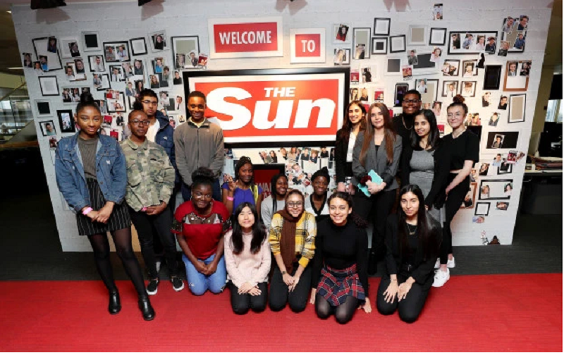17 aspiring journalists posing in front of a red and white sign that reads Welcome to The Sun. Eight of the young people are standing and nine are kneeling on red carpet.