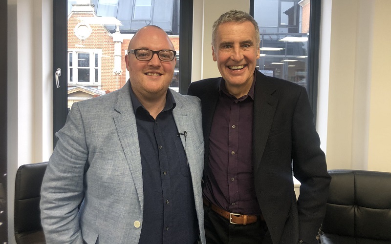 Dermot Murnaghan in a burgundy shirt and black jacket on the right standing with his arm around News Associates head of journalism Andrew Greaves in a dark blue shirt and light blue jacket. They both have big grins on their faces.