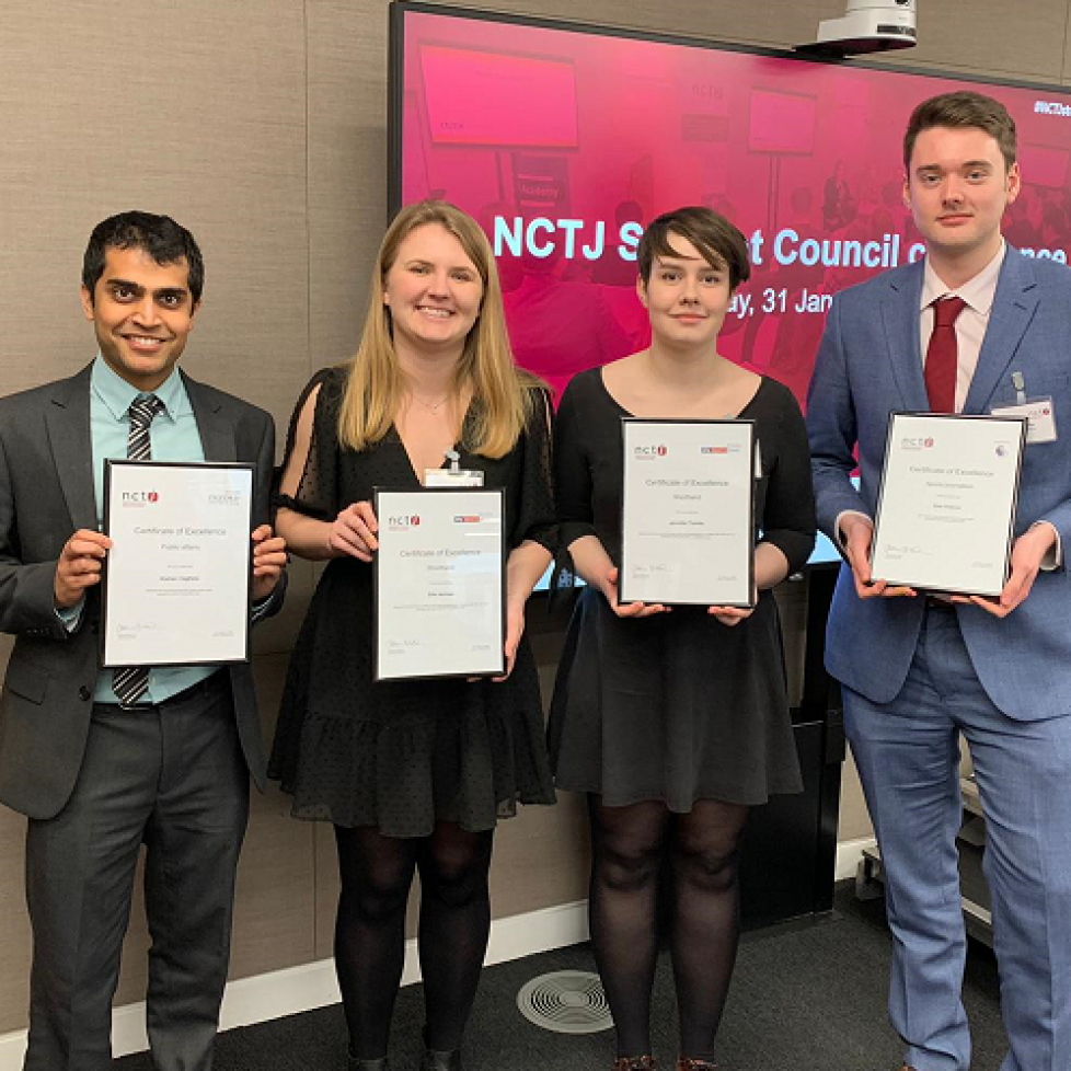 News Associates graduates Kish, Ella, Jen and Sam with their NCTJ diploma awards. All four are smartly dressed and standing in a row posing holding their framed certificates. Behind them is a pink sign advertising the event.