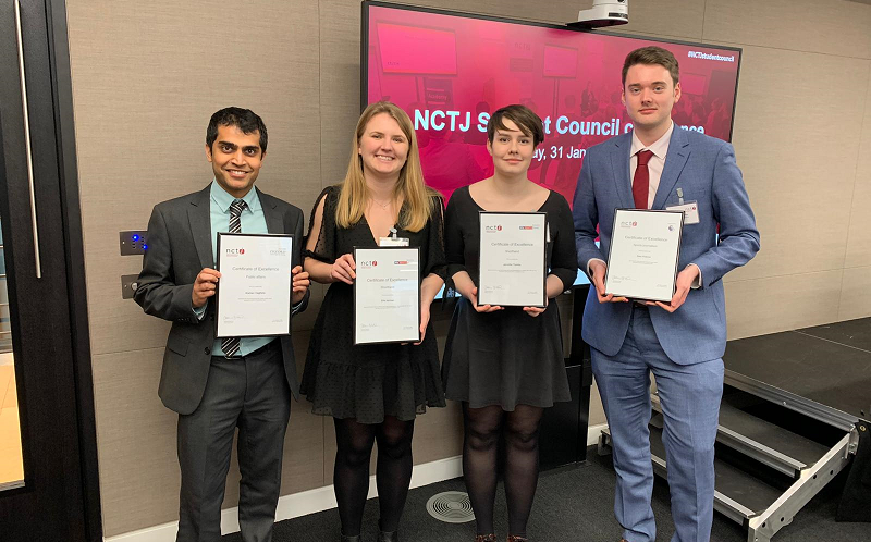 News Associates graduates Kish, Ella, Jen and Sam with their NCTJ diploma awards. All four are smartly dressed and standing in a row posing holding their framed certificates. Behind them is a pink sign advertising the event.