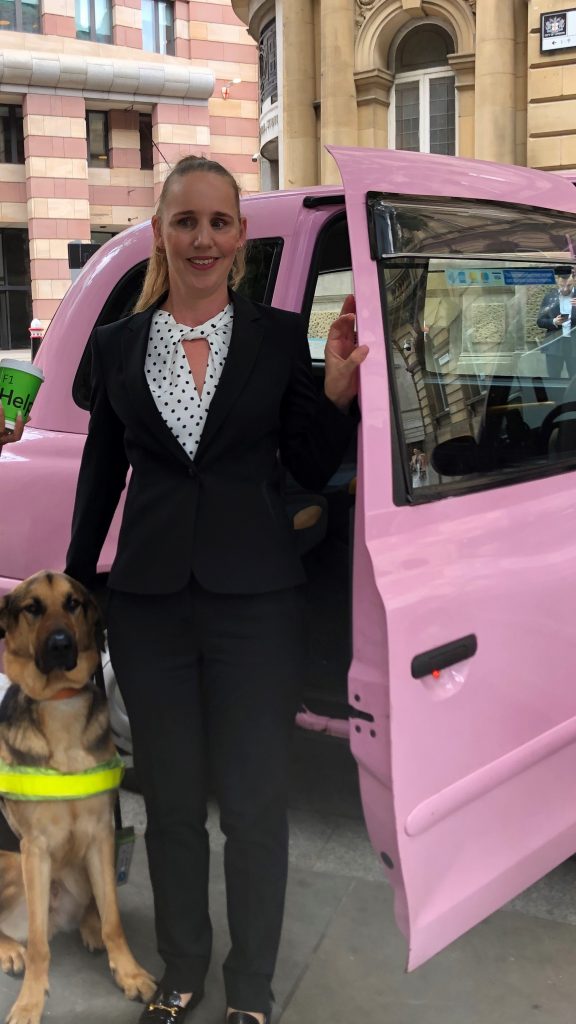 Kate Pounds with her blonde hair tied back wearing a white and black spotty blouse with a black jacket and black trousers standing next to a bright pink taxi next to her guide dog Bertie outside the Bloomberg offices.