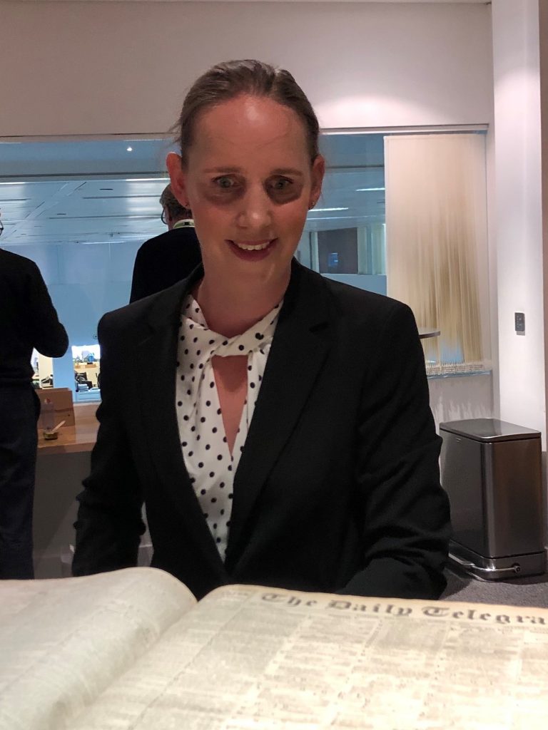 Kate Pounds with her hair tied back, wearing a white and black spotty blouse and a black jacket, looking a 100-year-old copy of The Daily Telegraph newspaper.