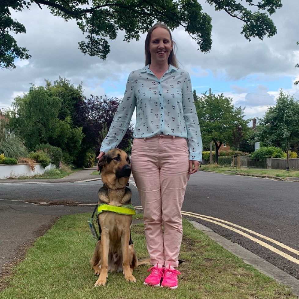 Kate Pounds and her guide dog Bertie outside on a patch of grass underneath a tree. Kate's blonde hair is down, she is wearing a blue blouse with pale pin trousers and pink trainers.