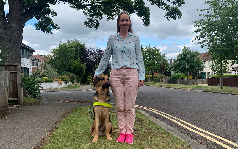 Kate Pounds and her guide dog Bertie outside on a patch of grass underneath a tree. Kate's blonde hair is down, she is wearing a blue blouse with pale pin trousers and pink trainers.