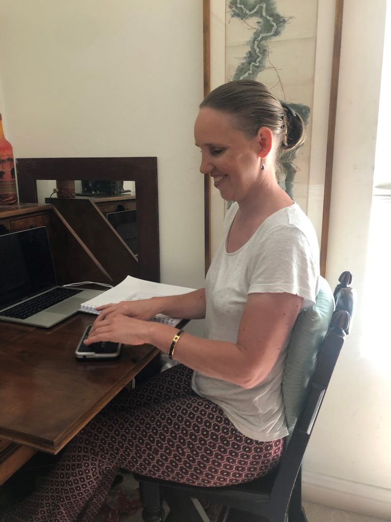 Kate Pounds looking very happy using her notetaker. She looks relaxed sitting at her mahogany desk on a mahogany chair with a green cushion behind her back. She is wearing a white t-shirt and pink patterned trousers. The notetaker is a small black device with keys. You can also see her Mac laptop and printed Braille code book on her desk.