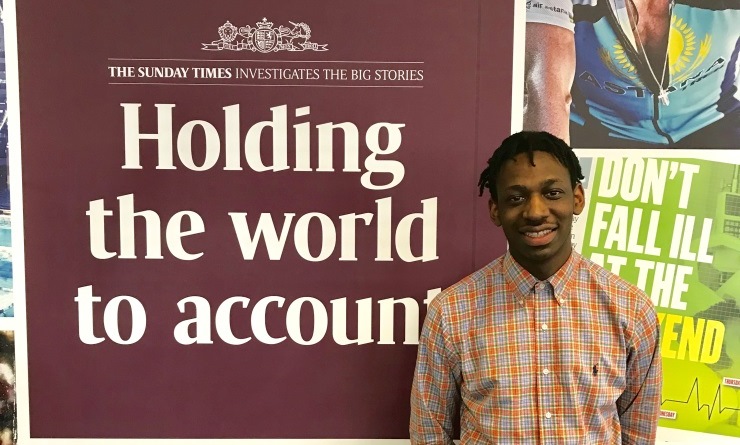 Shingi Mararike wearing an orange and blue checked shirt smiling, standing in front of a burgundy Sunday Times poster that reads 'Holding the world to account'.