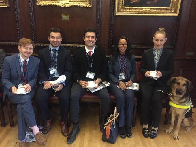 Five News Associates journalists sitting on dark wooden chairs with burgundy padding against a dark wooden wall at the Society of Editors' conference in Stationers’ Hall in London. They are all very smartly dressed. Left to right are Matt Banks, Joshua Graham, Will Cracknell, Sammy Mngqosini and Kate Pounds. To the right of Kate is her guide dog Bertie.