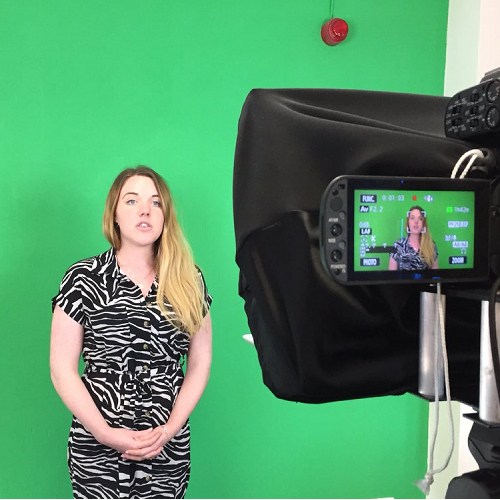 News Associates trainee Emily wearing a black and white jumpsuit presenting in front of a green screen wall being filmed by a black camera on a tripod. The image is to reflect the changes made to the diploma resulting in it becoming a level 5 qualification.