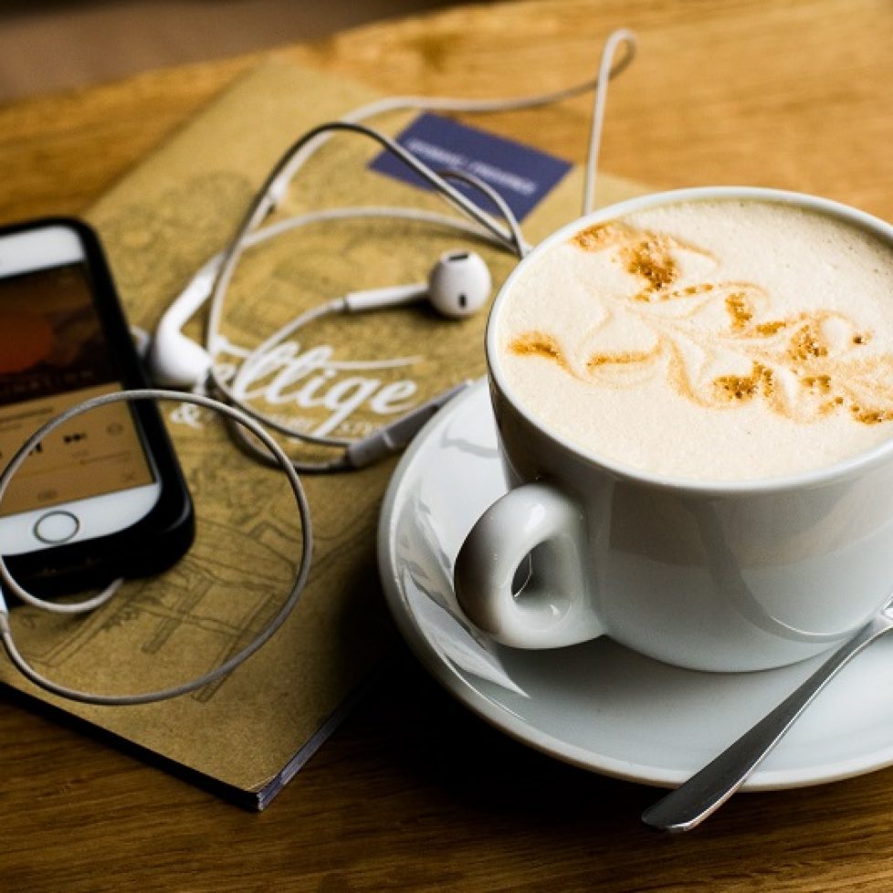 iPhone and coffee mug stock photo. White iphone is on the left, showing a podcast or song being played. White earphones are lying on top of a brown coffee shop menu, with a creamy latte in a mug on the right, sitting on a saucer with a teaspoon next to it. The objects are placed on a wooden table.