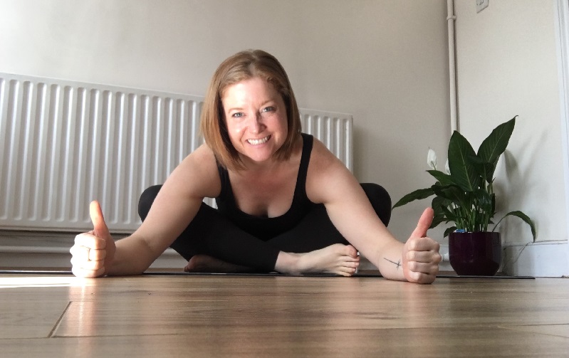 Yoga teacher Emma in a black strap top and black leggings. She is sitting at home on her wooden floor. Her legs are crossed and she is leaning forward with her arms stretched and her hands on the floor in a thumbs up. Emma has short brown and a huge grin. The white background is quite plain but to Emma's left is a luscious green leafy house plant in a dark pot. 