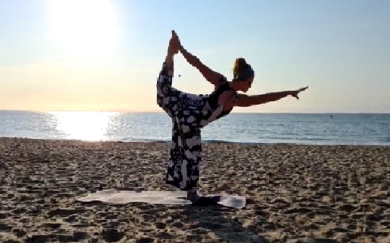 Yoga teacher Emma stretching on a sunny beach. The yellow sands, blue sea and blue sky are all idyllic and the sun is setting. Emma is right in the middle of the shot. She is wearing a black and white jumpsuit and her short brown hair is tied up. Her right arm is out in front of her, parallel to the ground. Her right leg is planted but she is holding her left leg up behind her with her left arm and her left knee is bent. It's a beautiful stand-up yoga pose.