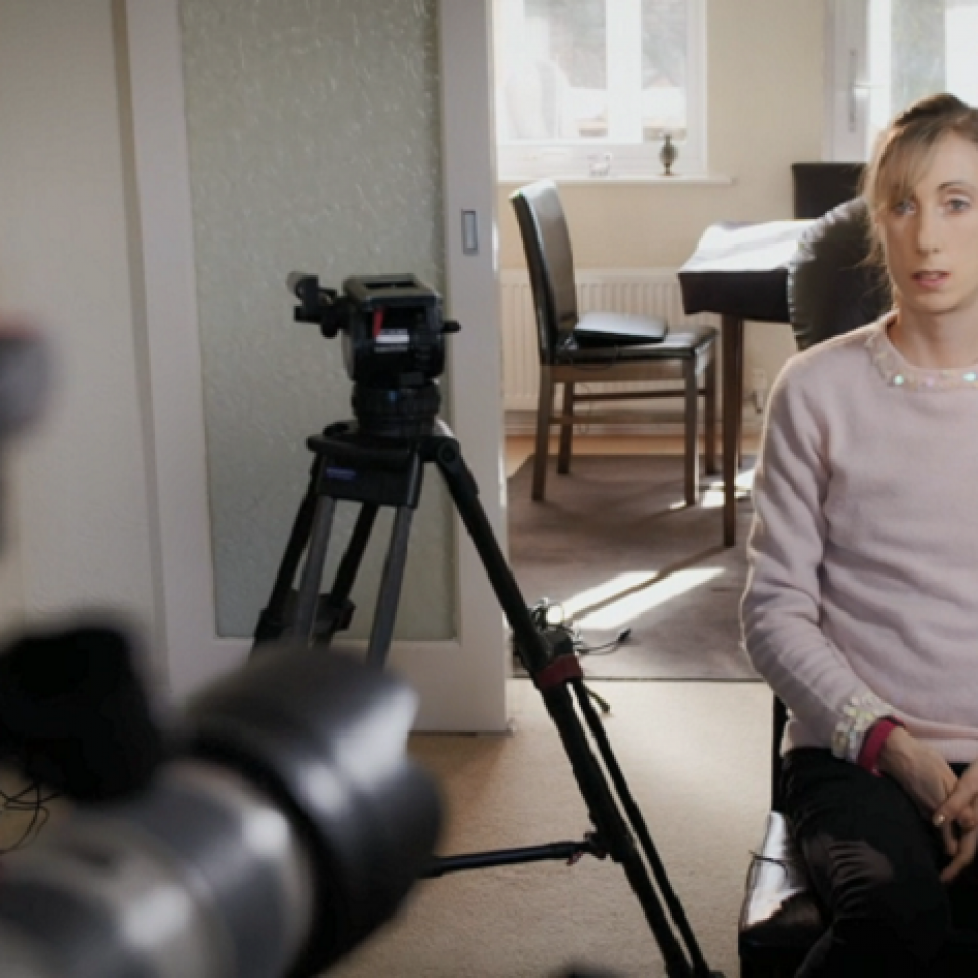 Screenshot from Panorama. Camera in foreground to the left, girl in pink jumper sitting on a chair to the right being filmed in a house.