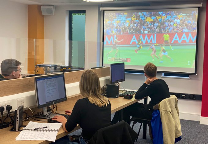 Three trainees sat socially distanced with a Perspex screen splitting the desk sitting in front of computers watching a screen showing Aussie Rules football.