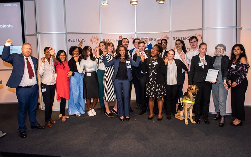 19 News Associates trainees and recipients of the Journalism Diversity Fund and our head of journalism Graham Moody at a JDF event at Reuters in 2019. Everyone is standing in a row, all dressed smart, cheering and smiling, many with their arms in the air. They are standing in front of a white wall with the black and orange Reuters loge printed on it.