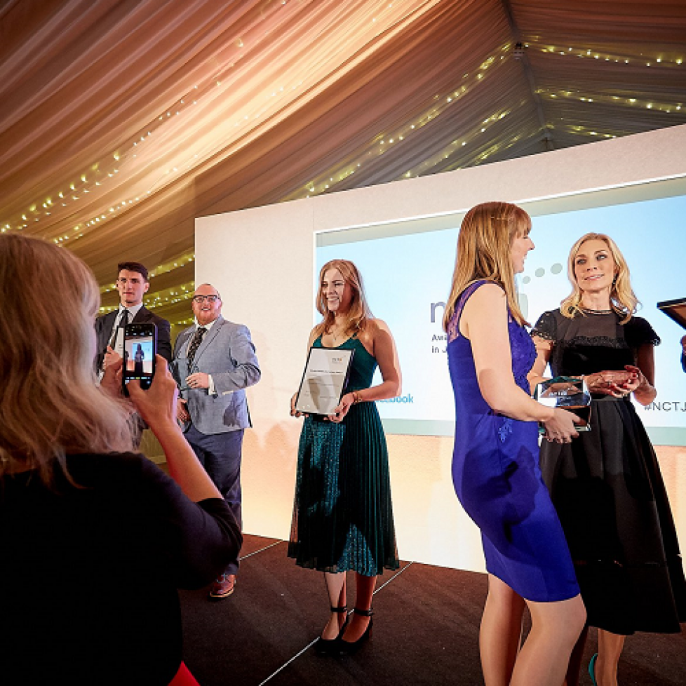 A candid shot of people on stage at the NCTJ Awards for Excellence in 2018. You can see graduate Jess Cripps in a beautiful emerald green sparkly dress posing for a picture holding her framed certificate which her mum is taking on an iphone which you can also see in shot. Other people, including head of journalism Andrew Greaves, are milling around.