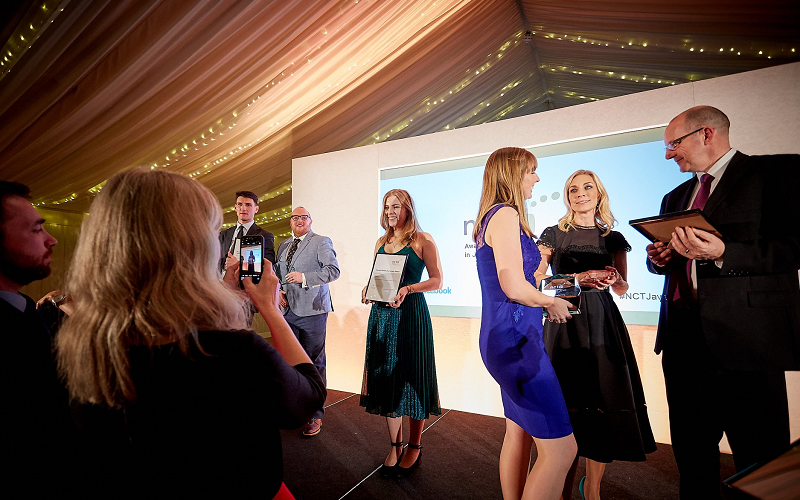 A candid shot of people on stage at the NCTJ Awards for Excellence in 2018. You can see graduate Jess Cripps in a beautiful emerald green sparkly dress posing for a picture holding her framed certificate which her mum is taking on an iphone which you can also see in shot. Other people, including head of journalism Andrew Greaves, are milling around.