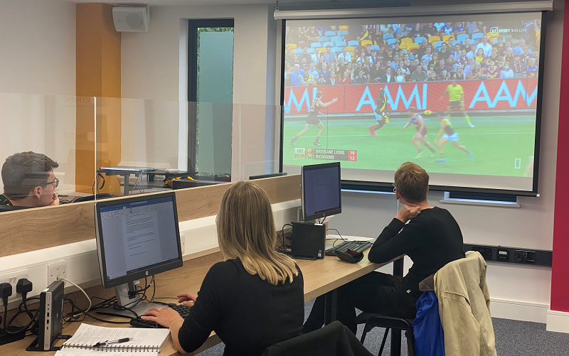 Three trainees on our journalism courses sat socially distanced with a Perspex screen splitting the desk sitting in front of computers watching a screen showing Aussie Rules football.