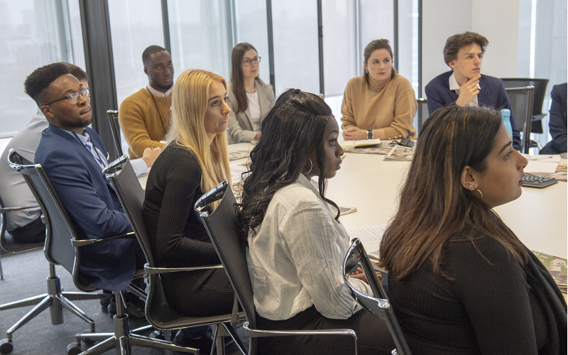 A group of News Associates/News UK trainees sitting around a large round table at News UK. You can see nine young journalists all dressed smartly looking at someone who is out of shot. 