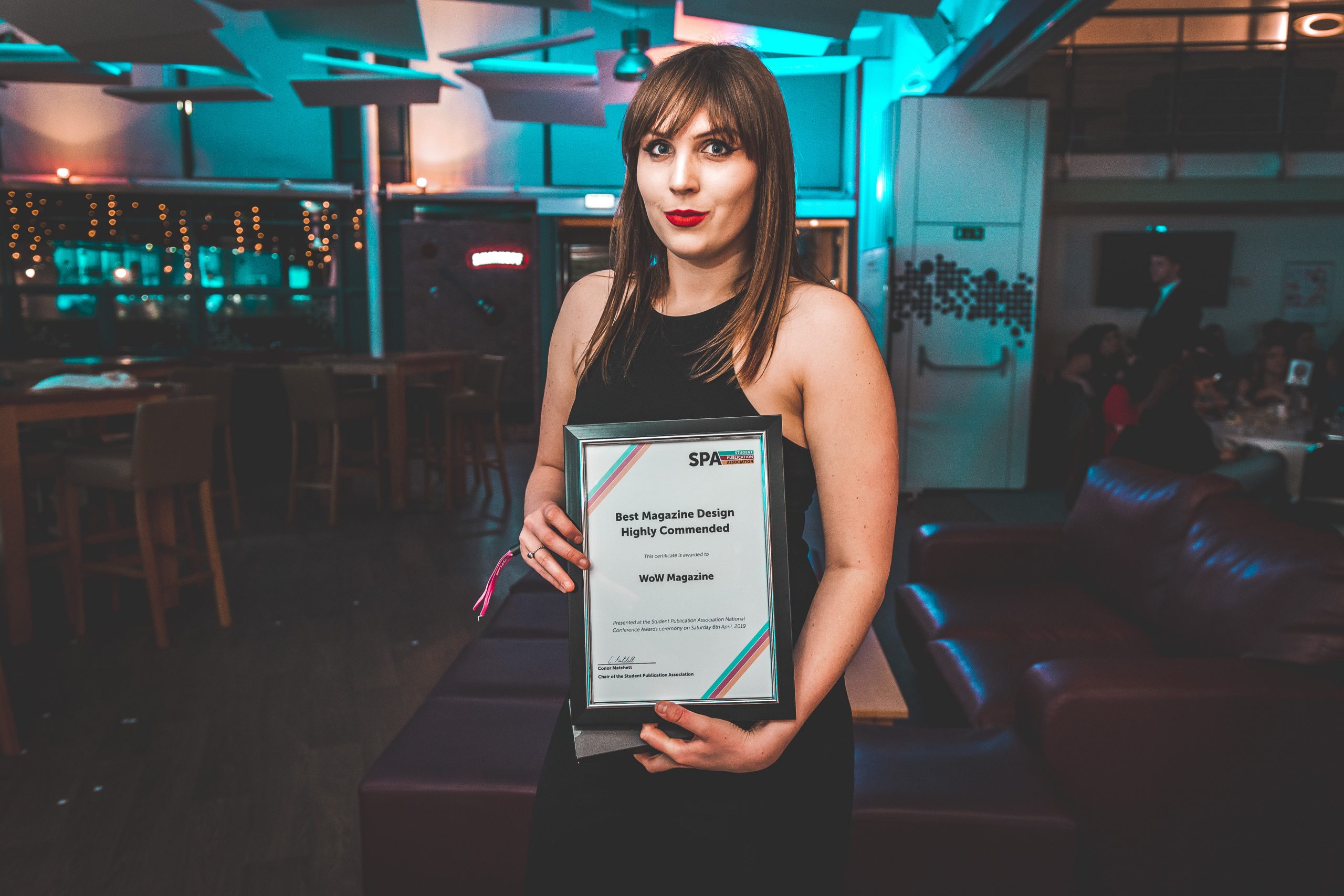 News Associates alumna Alice collecting with her award at the SPA National Conference in 2019. She's holding her framed certificate posing at the camera. She's wearing a formal black dress, her brown hair is straight and she's wearing red lipstick. 