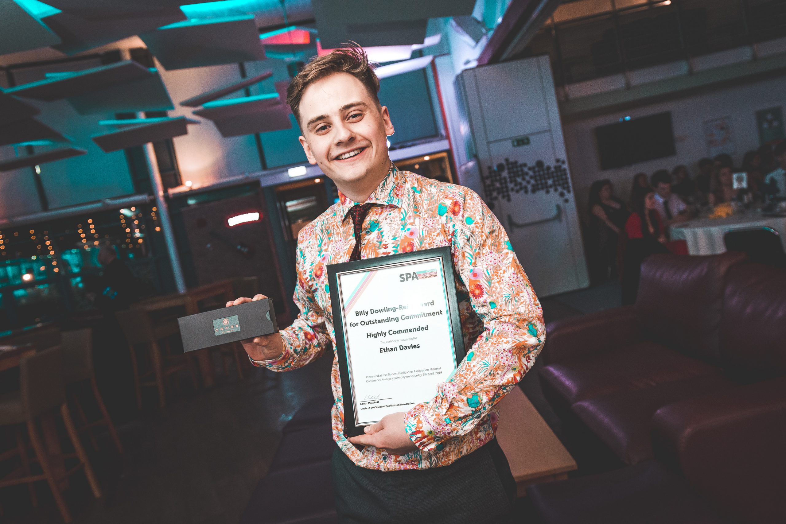 News Associates alumni Ethan with her award at the SPA National Conference in 2019. 2019. He's holding her framed certificate posing at the camera. He's wearing a very jazzy orange and turquoise floral shirt and has a huge grin. 