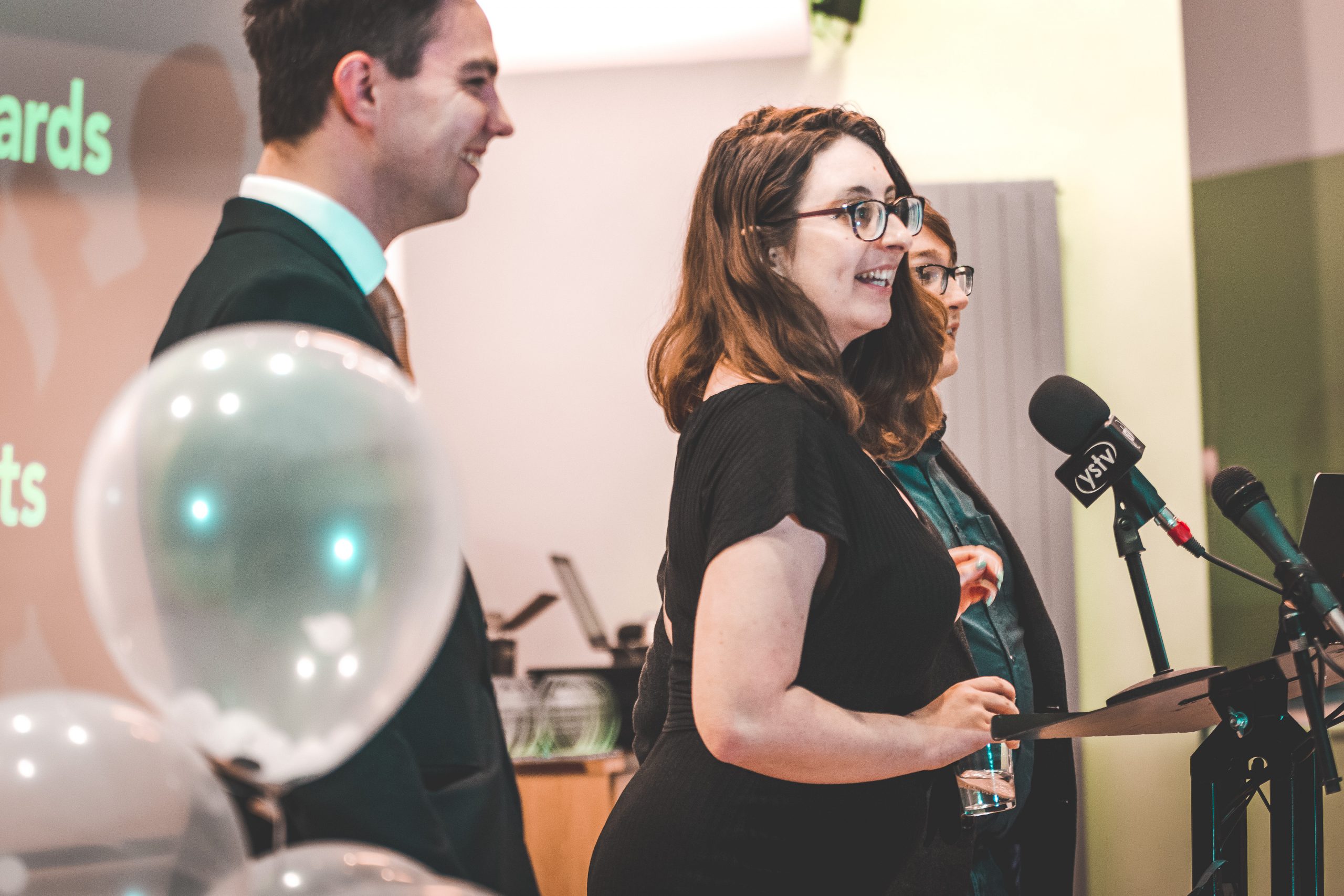 Jem Collins presenting an award at the SPA National Conference in 2019. It's a side angle as she talks into a microphone on a podium. She's dressed in all black and her dark brown hair is down. She's wearing glasses and holding a glass of water. She's smiling but it's a candid shot.