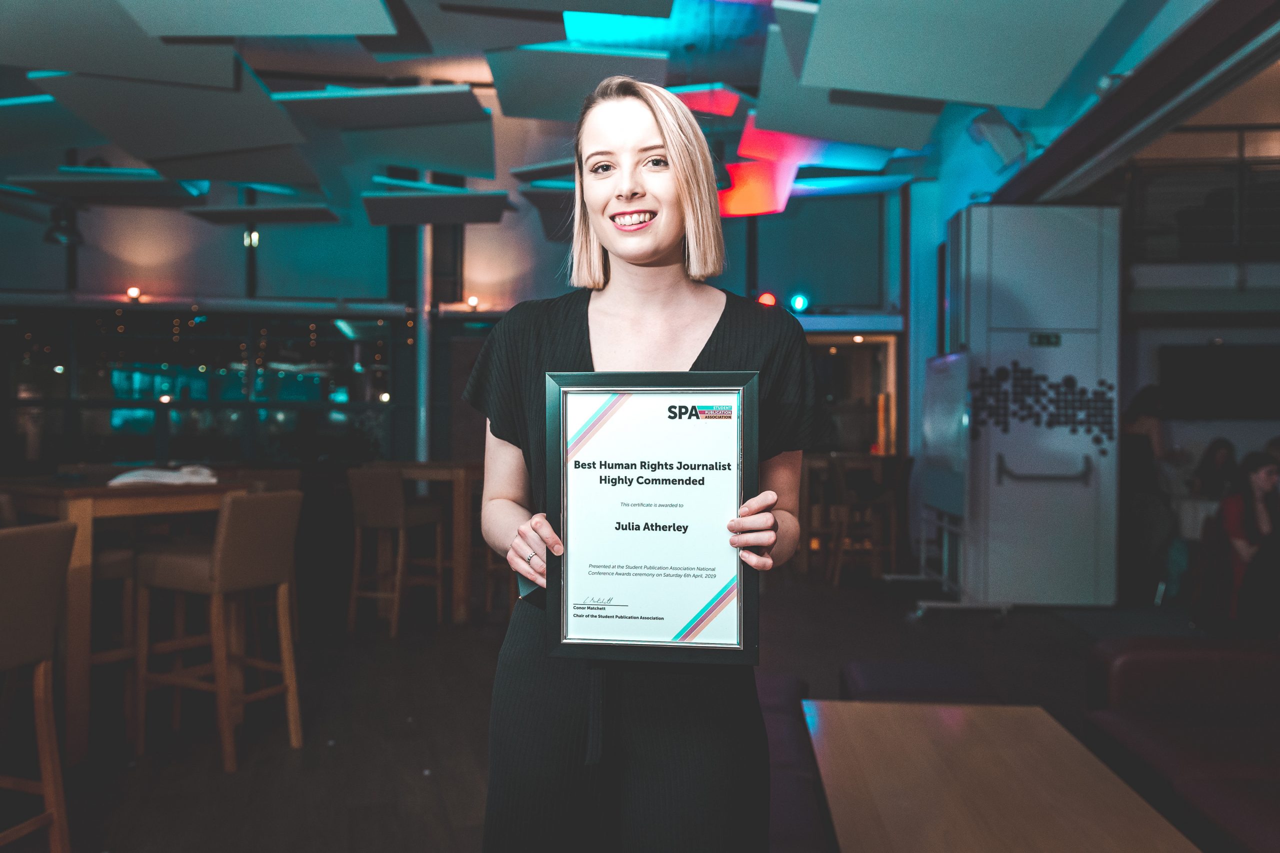 News Associates alumna Julia with her award at the SPA National Conference in 2019. 2019. She's holding her framed certificate posing at the camera. She's wearing a formal black dress, her blonde hair is straight and she's smiling. 