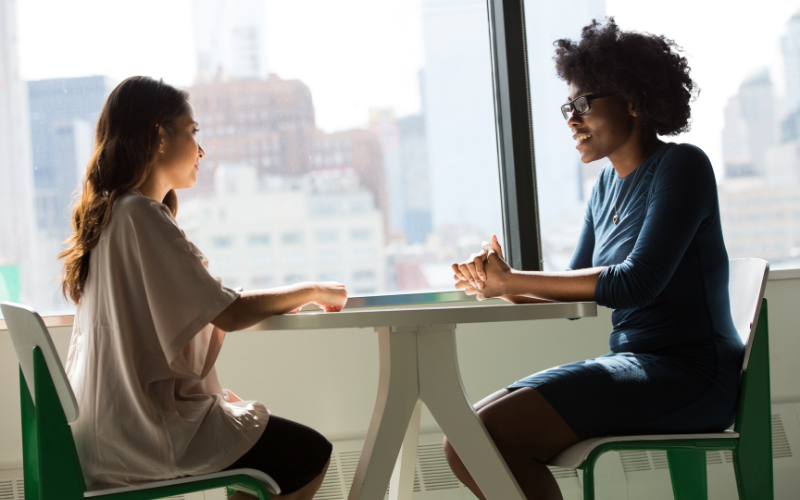 Stock image of two women talking at an office table. Both women have their hands on the table and are smiling, and there is a big window in the background.
