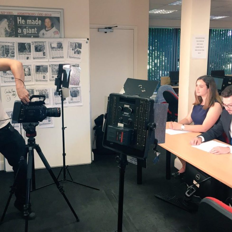 Trainees filming a news bulletin, cameraperson on the left and two presenters sat on the right. Photo taken in newsroom/classroom