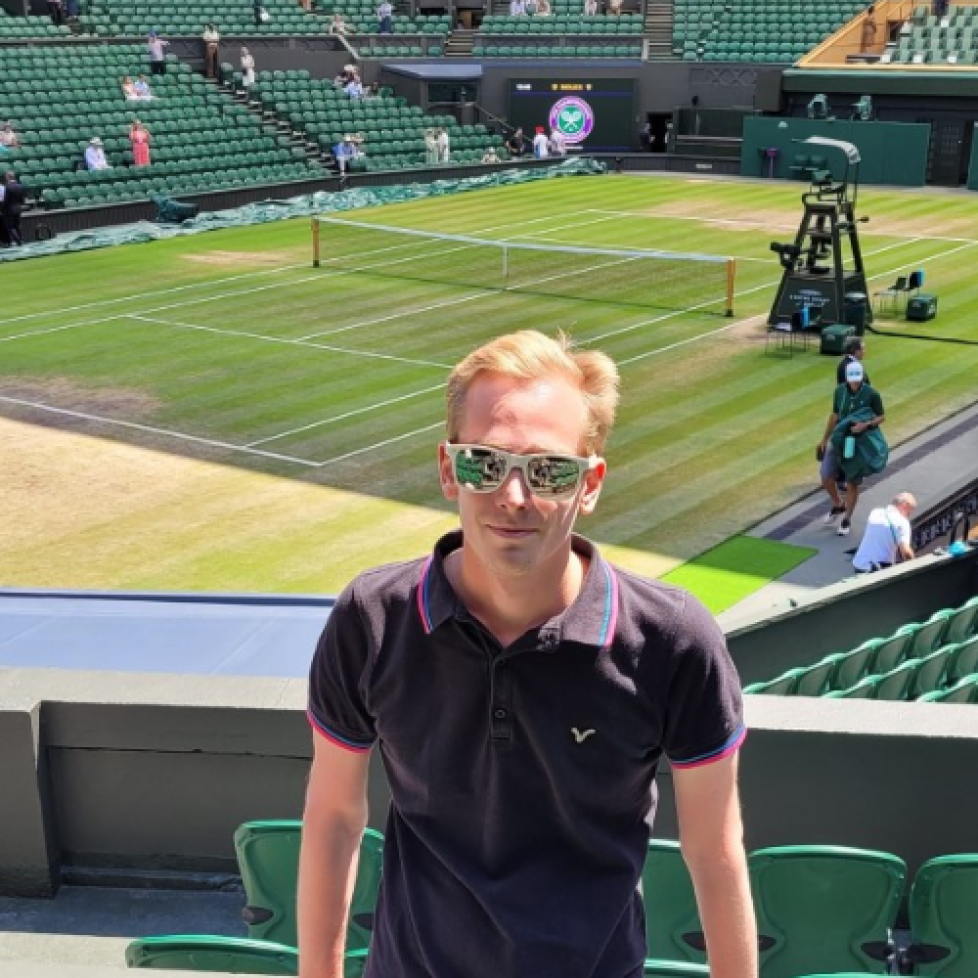 Photo of Oli standing at Wimbledon with court behind him. He is wearing a dark t-shirt and sunglasses.