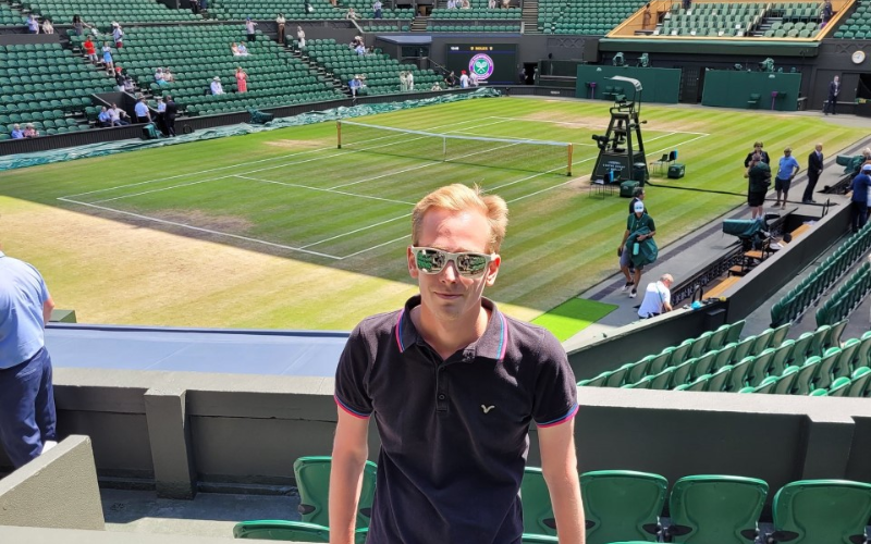 Photo of Oli standing at Wimbledon with court behind him. He is wearing a dark t-shirt and sunglasses.