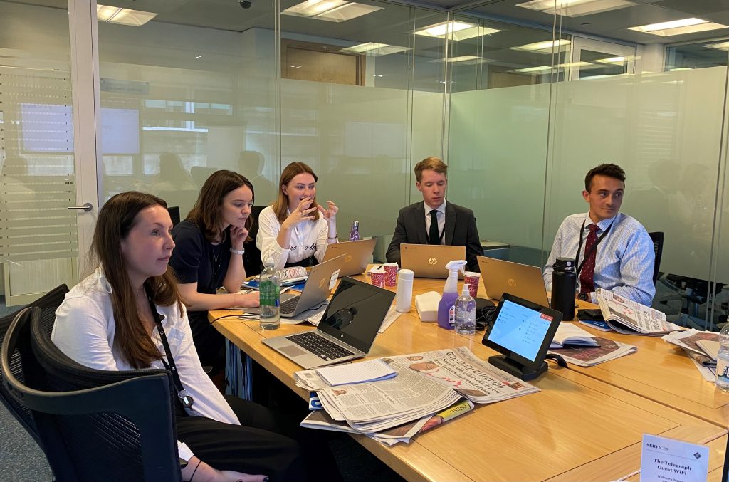 Five Telegraph editorial graduates around a desk with laptops and newspapers.