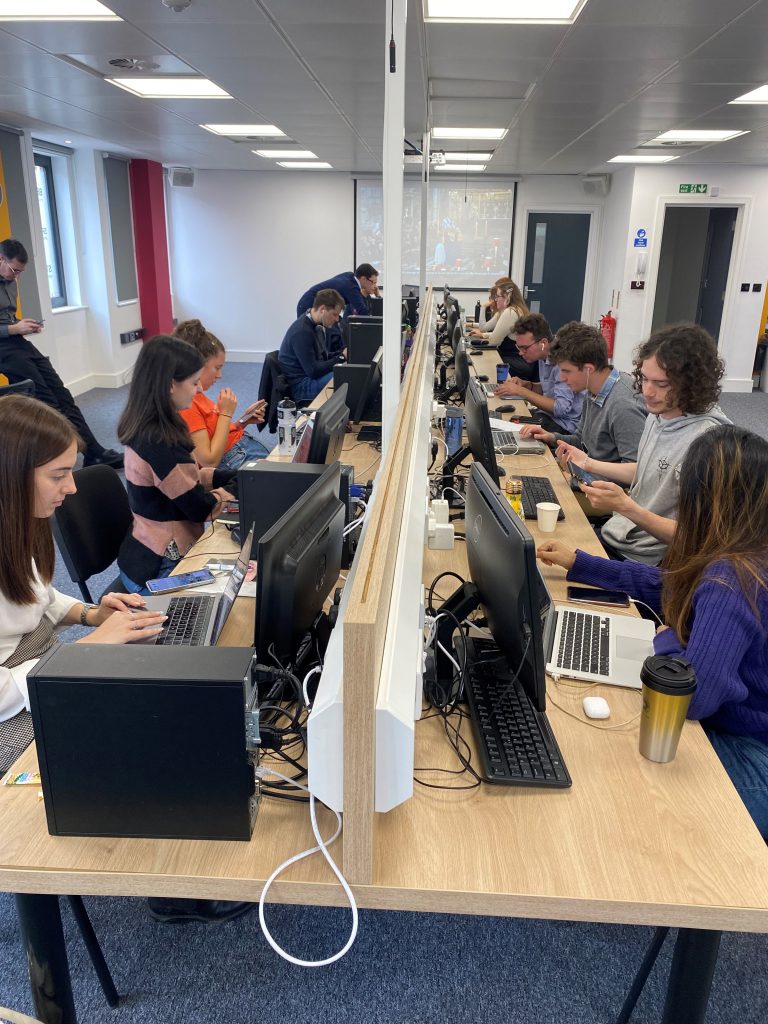 A desk of News Associates trainees working on computers and laptops with a screen showing the news in the background.