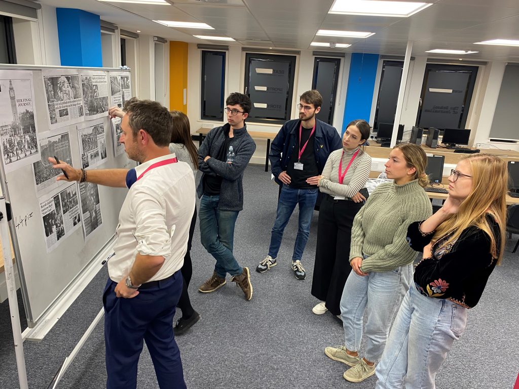 A group of five trainees on The Times graduate scheme standing looking at a whiteboard with drafts of their 16-page e-edition stuck to the front. News Associates managing editor James Toney is standing in front of them talking through the pages. 