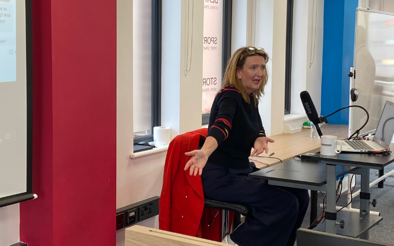 Victoria Derbyshire is sitting on a chair at the front of a classroom in front of a window and talking to journalism students. 