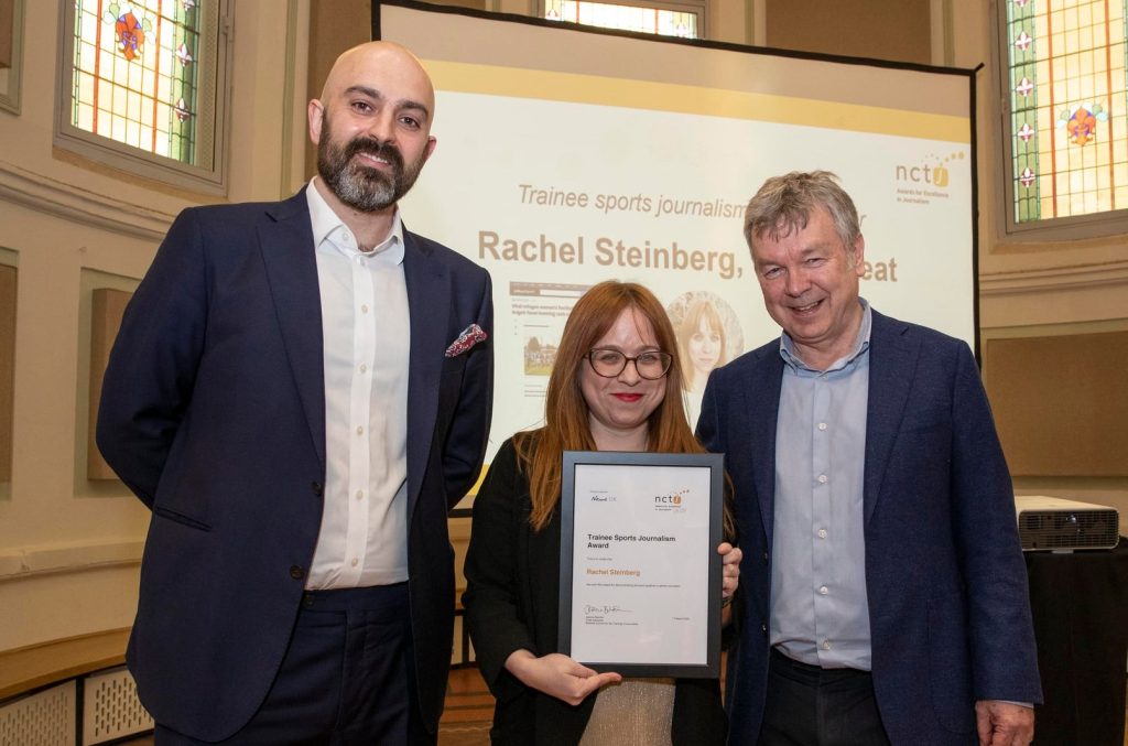 Rachel Steinberg collecting her trainee sports journalism award from Mark Hudson and Nick Powell in Manchester. Rachel is standing in the middle holding her framed A4 certificate with a big smile on her face. Mark is on her left and Nick on the right, they are both wearing suits and smiling for the camera. 