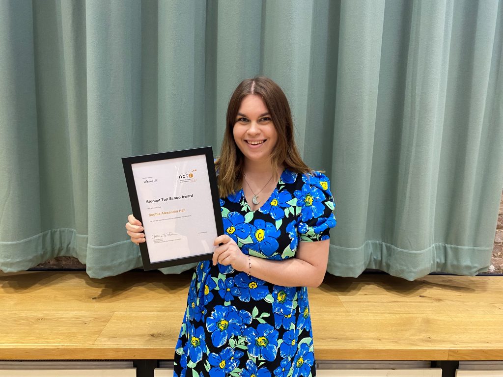 Sophia Alexandra Hall with her student top scoop award at the NCTJ Awards for Excellence 2021. Sophia is smiling holding her A4 framed certificate in a blue floral dress in front of a light green curtain.