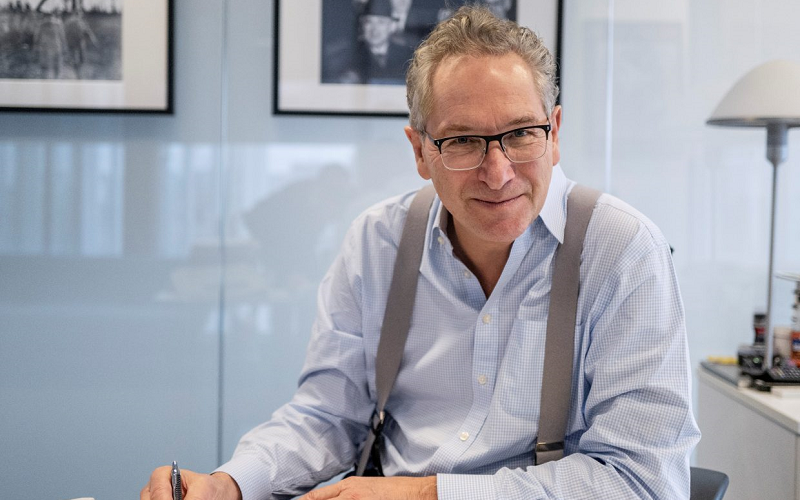 Times Newspapers chairman John Witherow sitting at a desk wearing a light blue shirt, smiling for the camera while holding a pen writing on paperwork. John Witherow is in a nice and bright glass office, behind him you can see a lamp and two pictures hung on the wall. We're using the image to illustrate he is the keynote speaker at our journalism conference JournoFest in 2023.