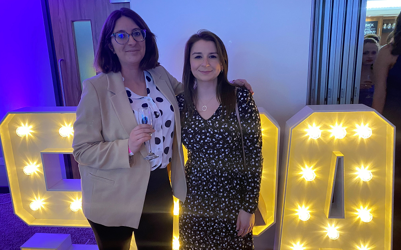 Jem Collins and Lucy Dyer at the Student Publication Association National Conference in Sheffield in 20233. Both are smartly dressed and standing smiling for the camera. They are standing in front of large light up letters spelling SPA.