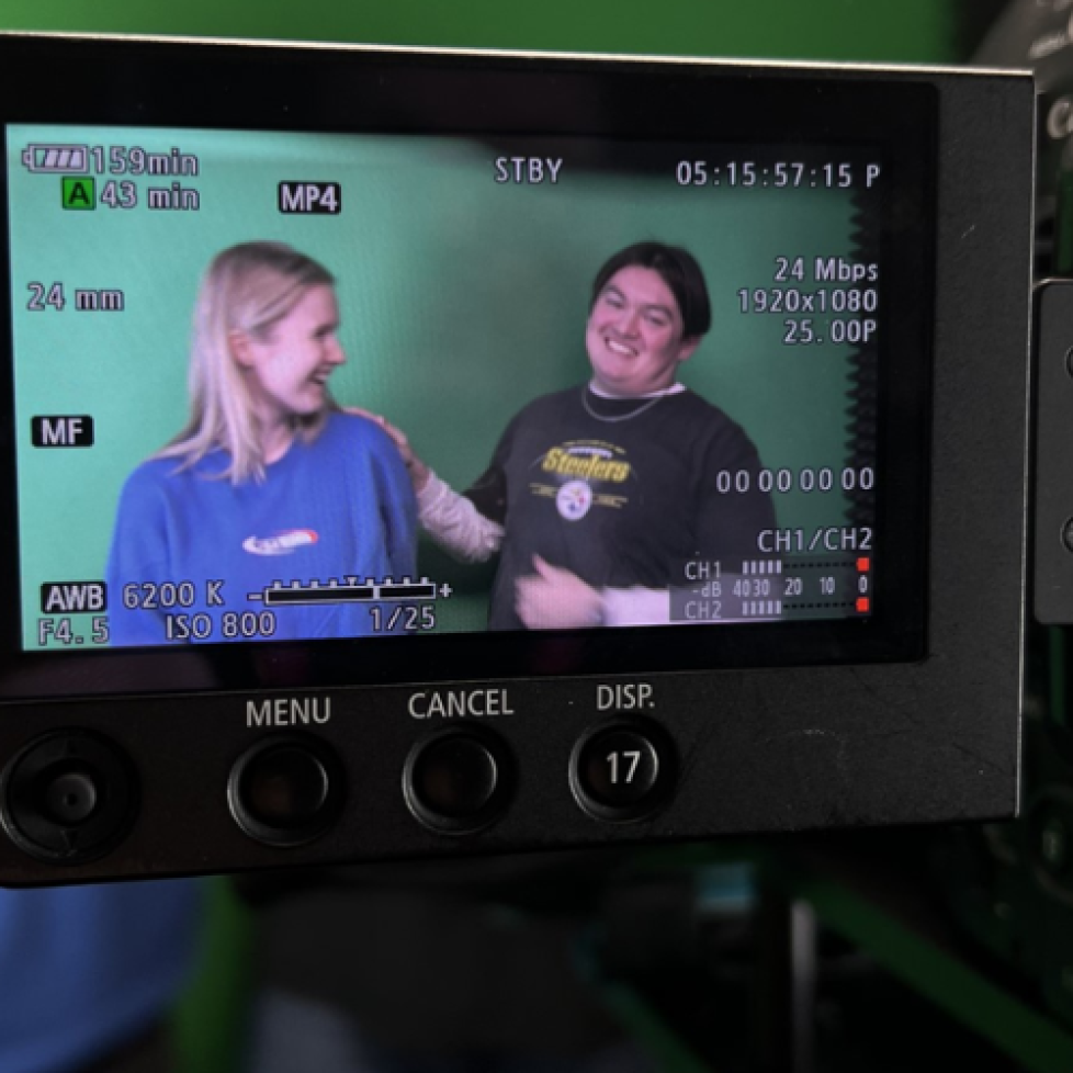 A photo of a camera lens showing two of our sports journalism trainees laughing in front of a green screen.