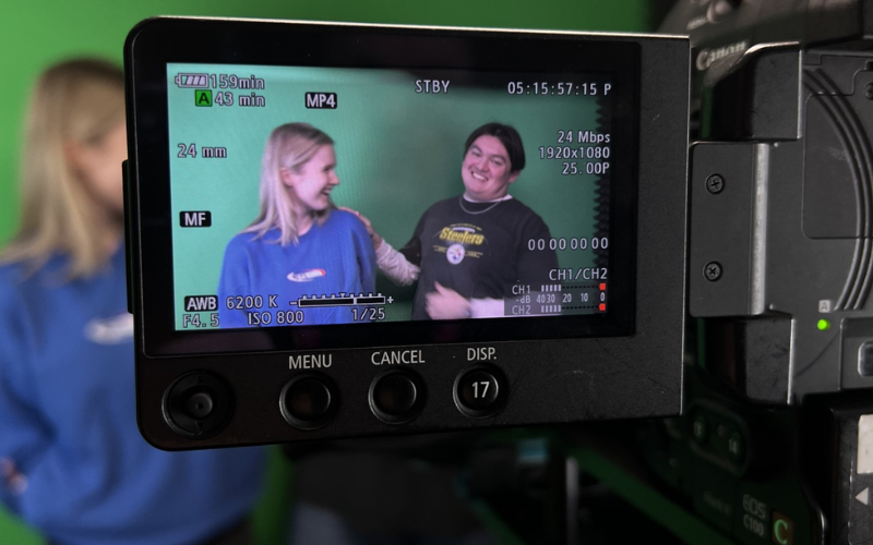 A photo of a camera lens showing two people laughing in front of a green screen. 