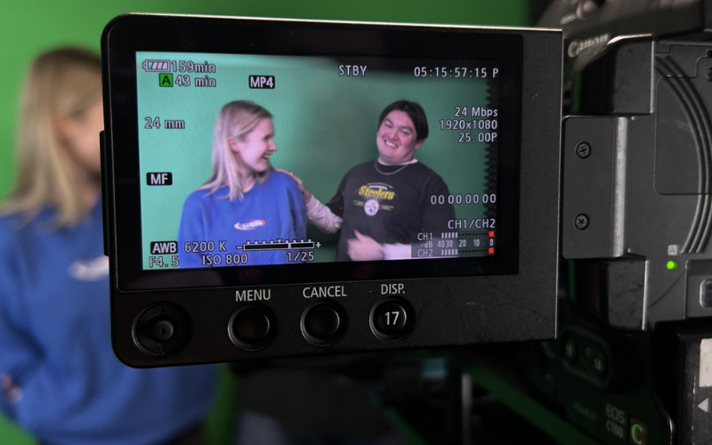 A photo of a camera lens showing two people laughing in front of a green screen.