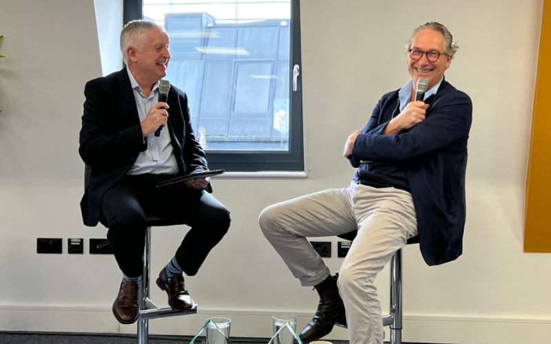 Graham Dudman and John Witherow sat on chairs holding microphones. They are laughing.