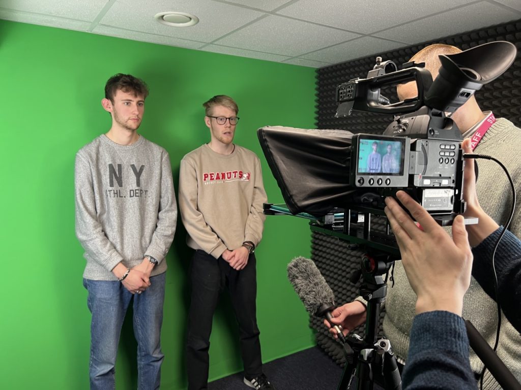 A photo of our two News Associates trainees standing in front of a green screen. They are standing in front of a camera. Someone is holding a microphone to the right of the photo. 