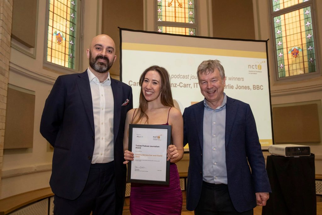 Carolina collecting her award for podcast of the year at the NCTJ Awards for Excellence 2022. Carolina is standing and posing holding her framed certificate with Mark Hudson and Nick Powell. They are all dressed smartly and are smiling.