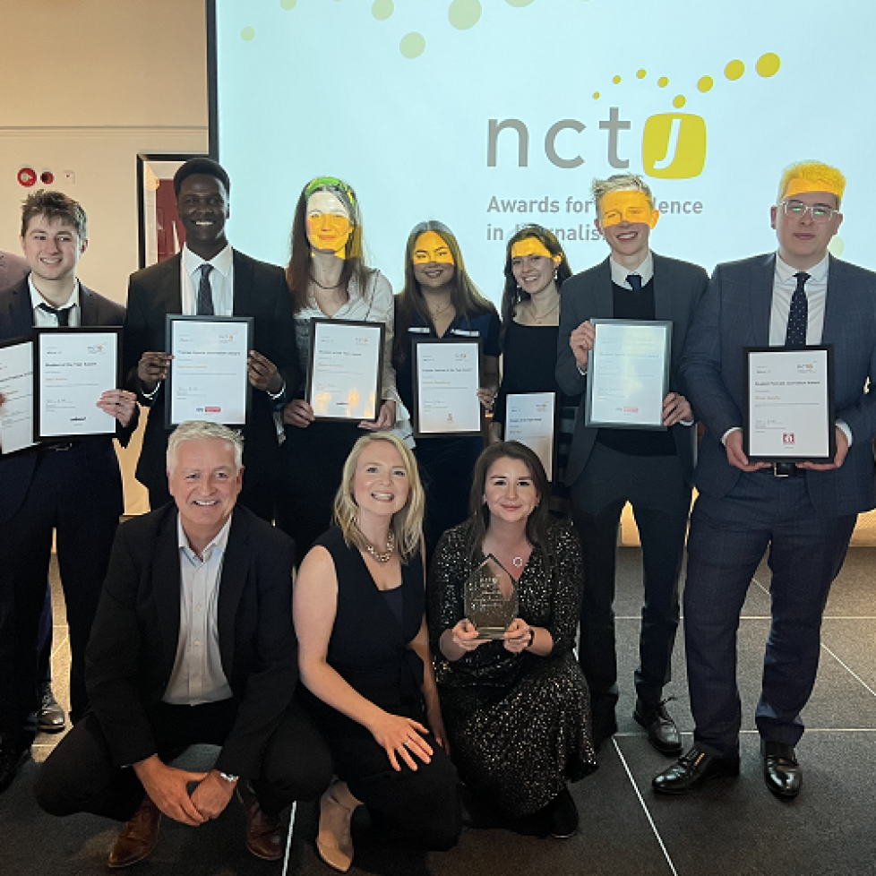 A group photo of all the News Associates winners and runners up at the NCTJ Awards for Excellence 2022. There are two rows of people, the back row of people are standing and the front row are kneeling. Everyone is dressed really smartly and are smiling for the camera. Some people are holding awards and framed certificates. There is a projector screen displaying the NCTJ logo behind everyone. The light from the projector is shining in some people's eyes!