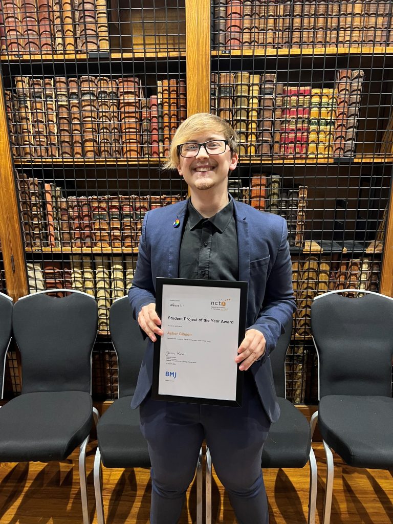 Asher Gibson with a massive smile holding his certificate. He is smartly dressed and standing in front of rows of books on a shelf in the library at the Royal College of Physicians at the NCTJ Awards for Excellence 2023.