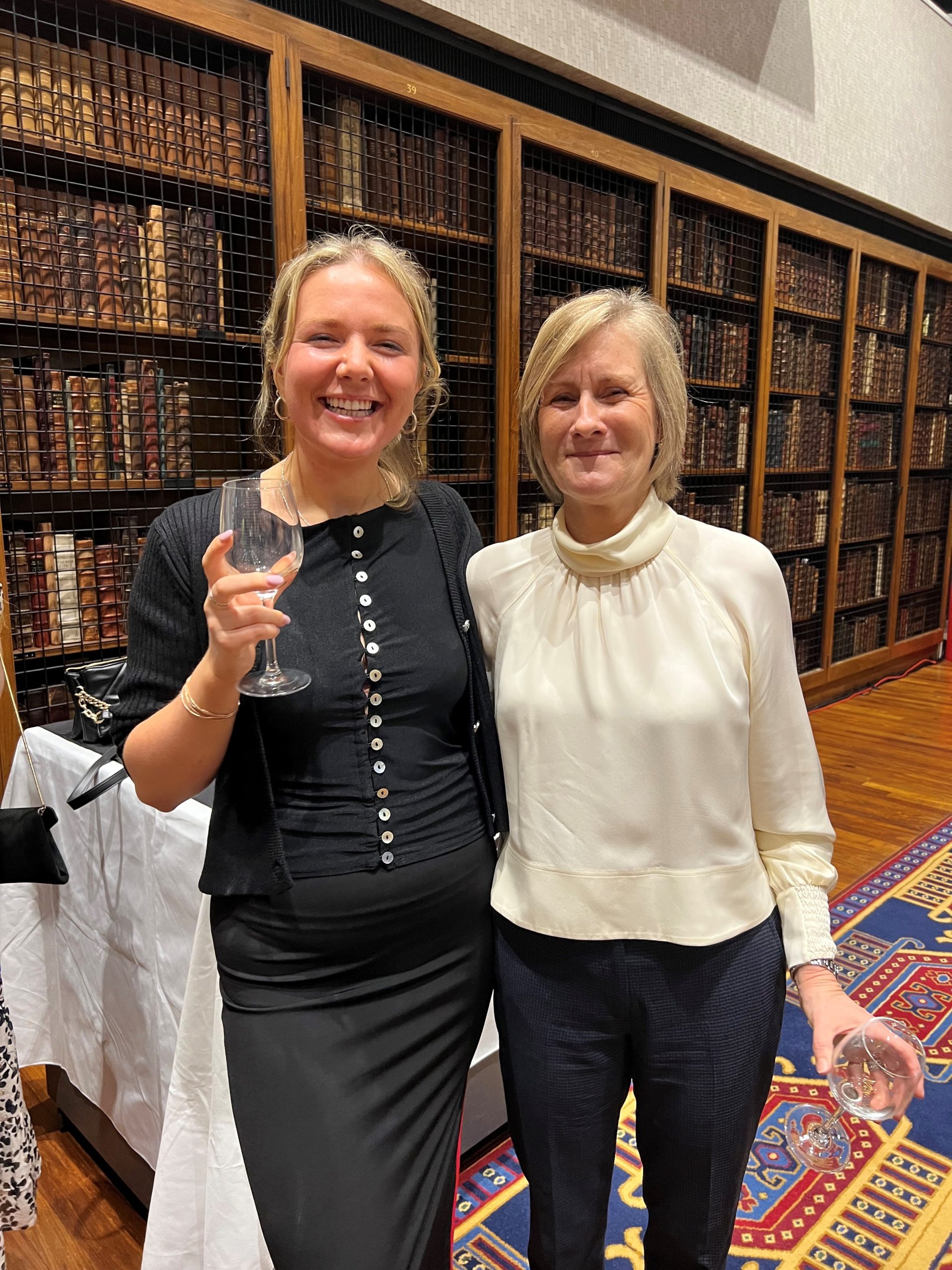 News Associates graduate Connie Bowker and shorthand tutor Angela Catto at the NCTJ Awards for Excellence 2023. They are both smiling and smartly dressed and standing in front of rows of books on a shelf in the library at the Royal College of Physicians.